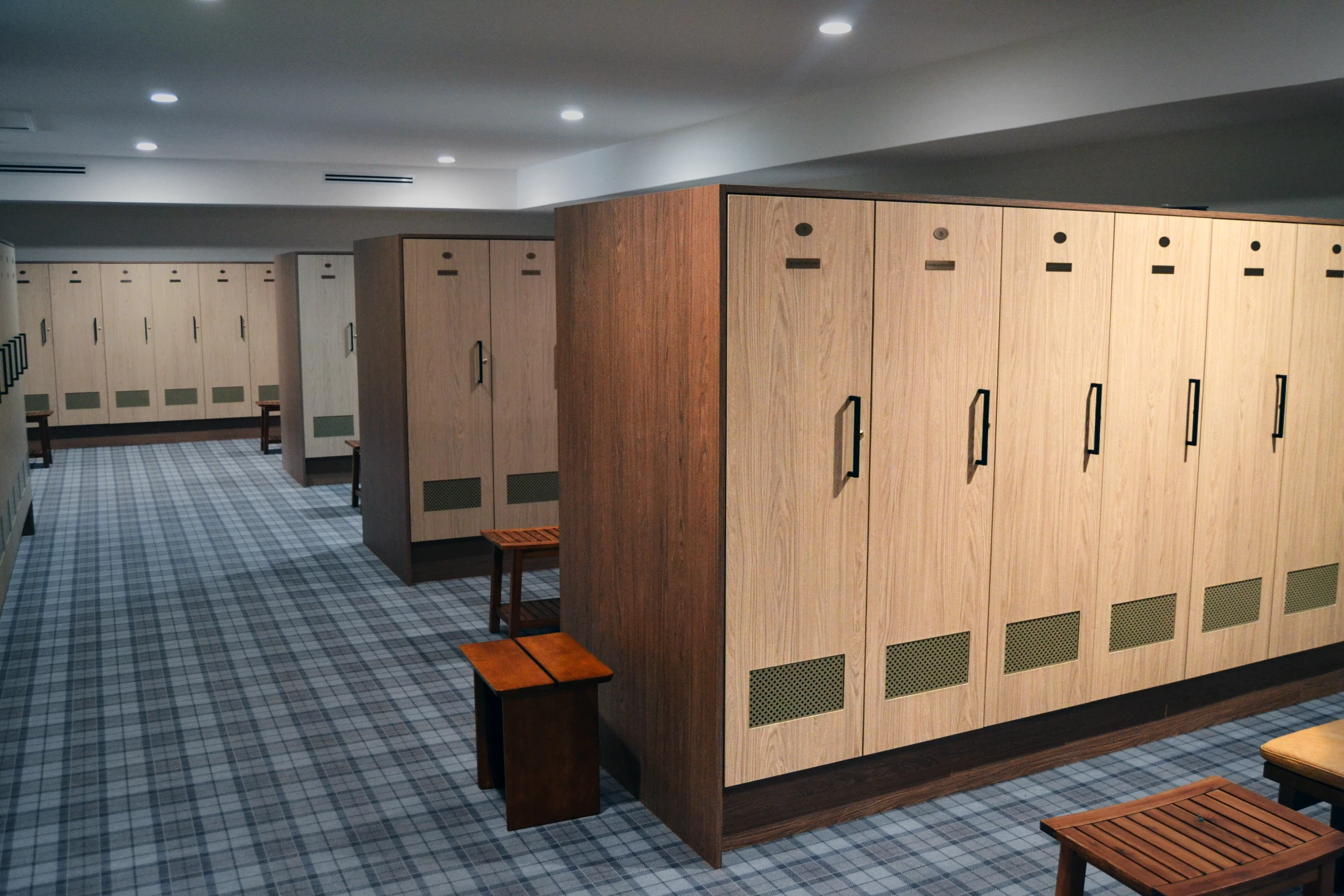 Private country club locker room with light oak lockers and green mesh ventilation panels, wood benches, and recessed ceiling lights.